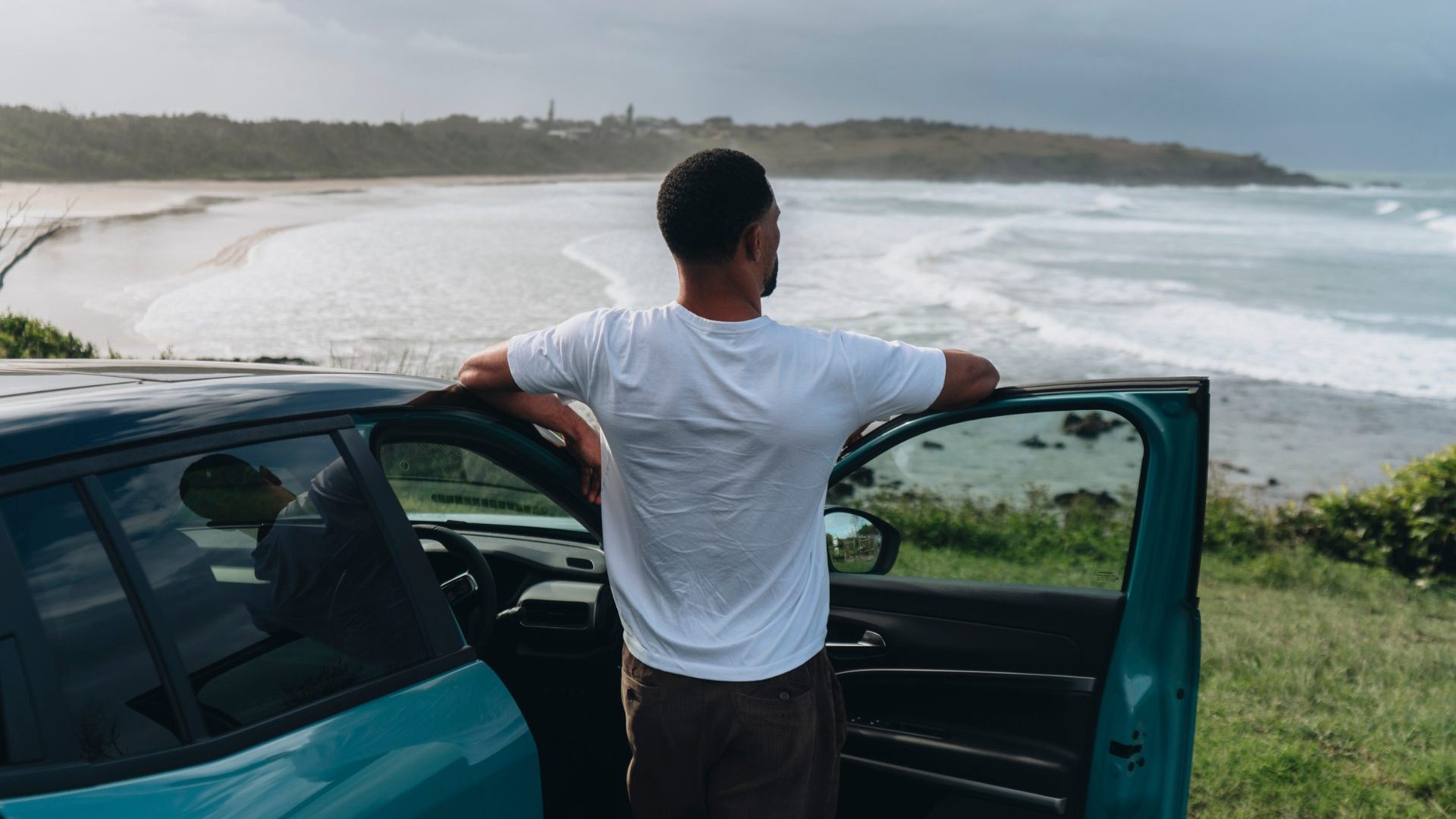 Man stands next to blue car overlooking surf