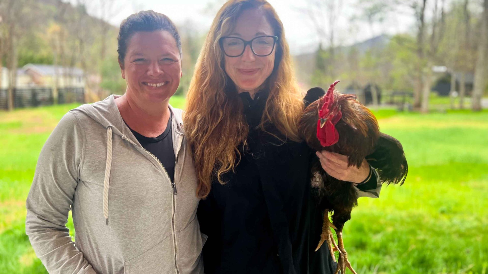 Two women, one with short dark hair, the other with long hair and glasses, smile to camera while holding a rooster.
