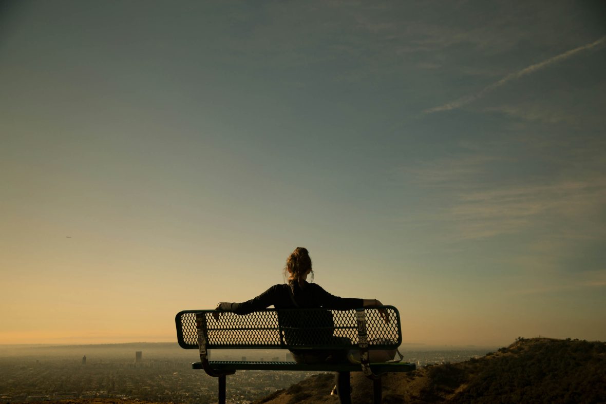 A woman sits on a bench and looks out at dusk over a city.