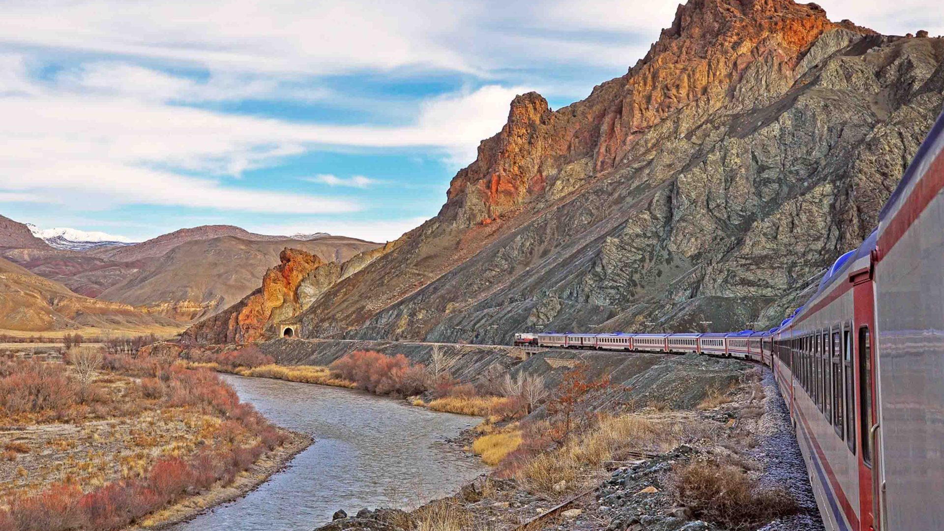 A train winds through the mountains, past a river, before heading under a mountain pass.