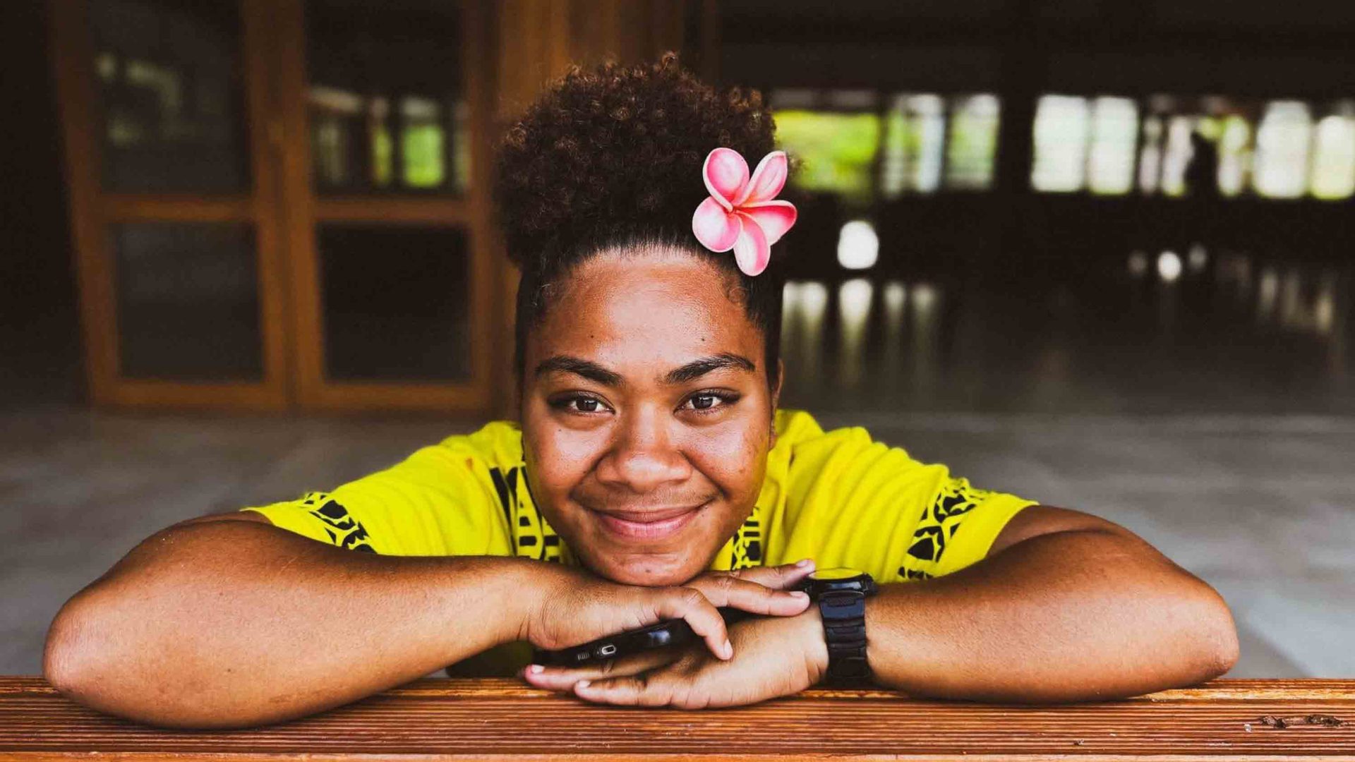 A woman with a yellow top and a flower in her hair, smiles to camera.