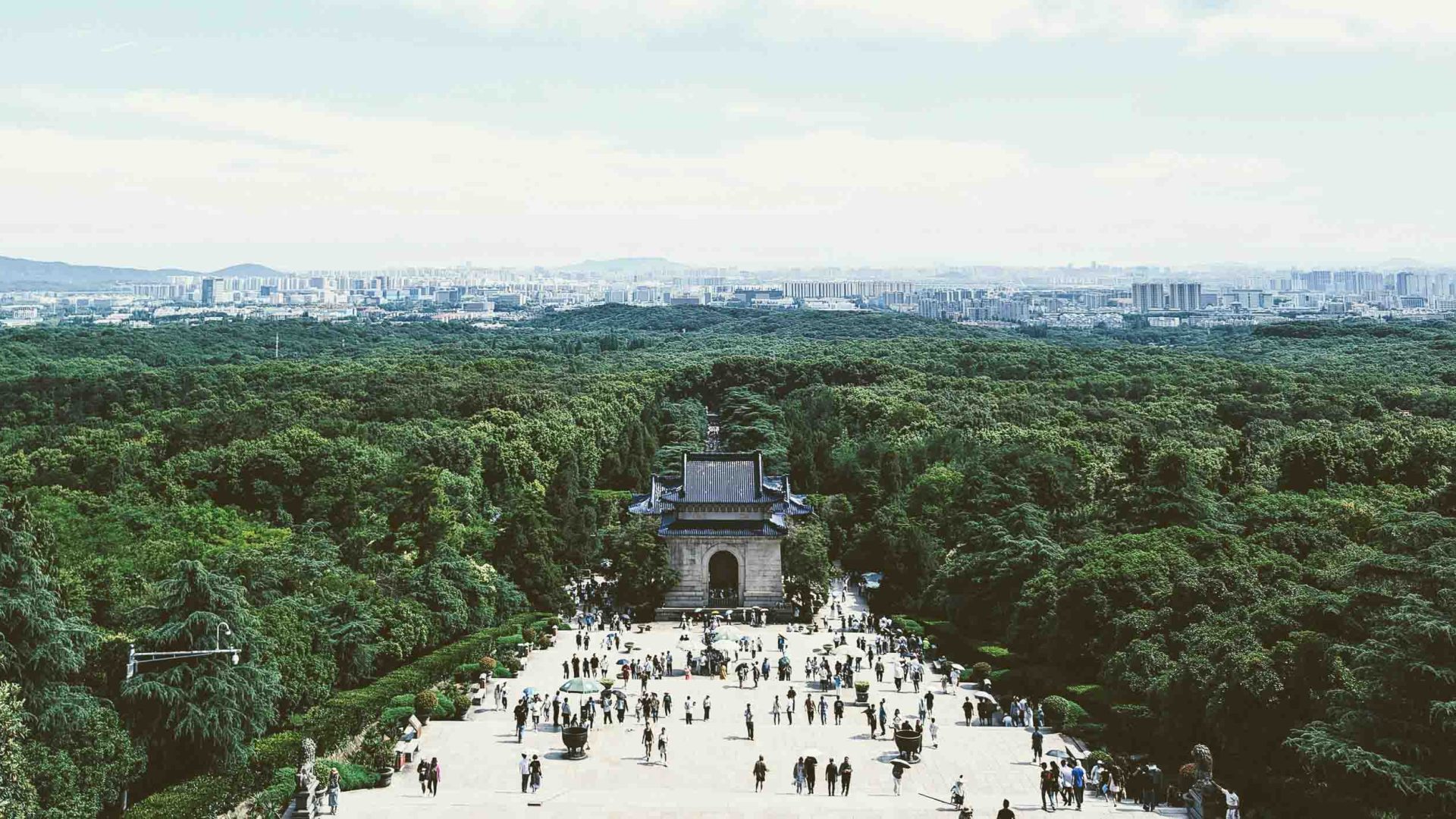 People walk toward a mausoleum in a forested area with a city in the background.