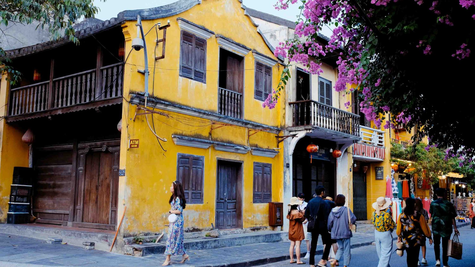 People walk past a mustard coloured building framed by bougainvillaea.
