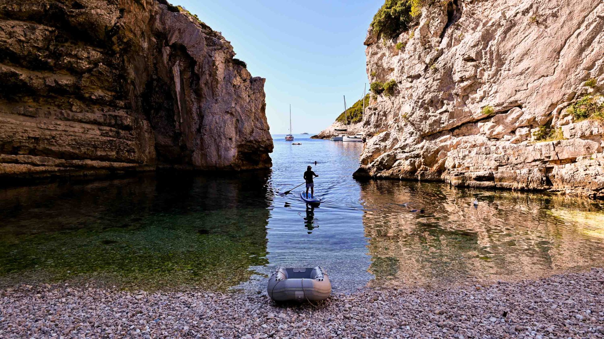 A boat rests on the shore and a person does stand up paddle in a calm bay.