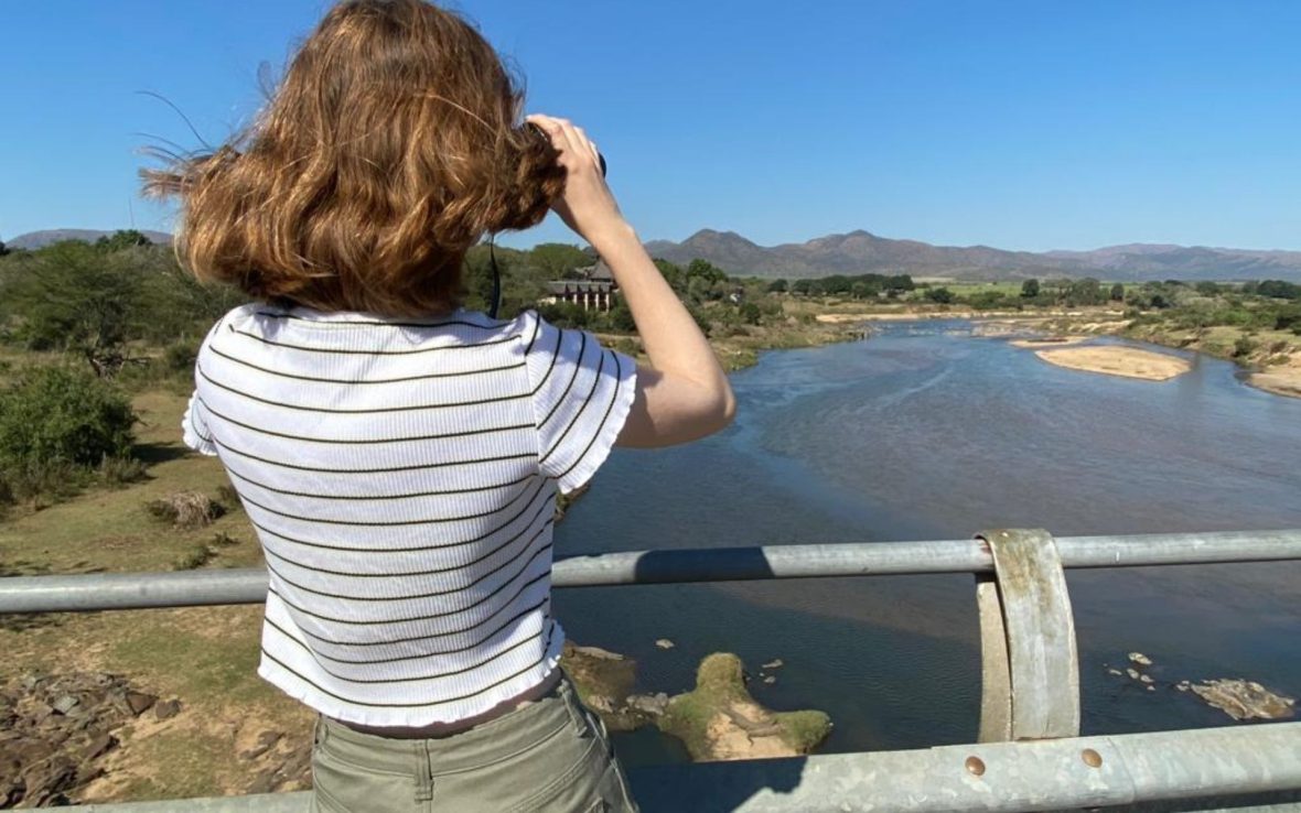 A woman on a bridge looks through binoculars at the river and mountains ahead