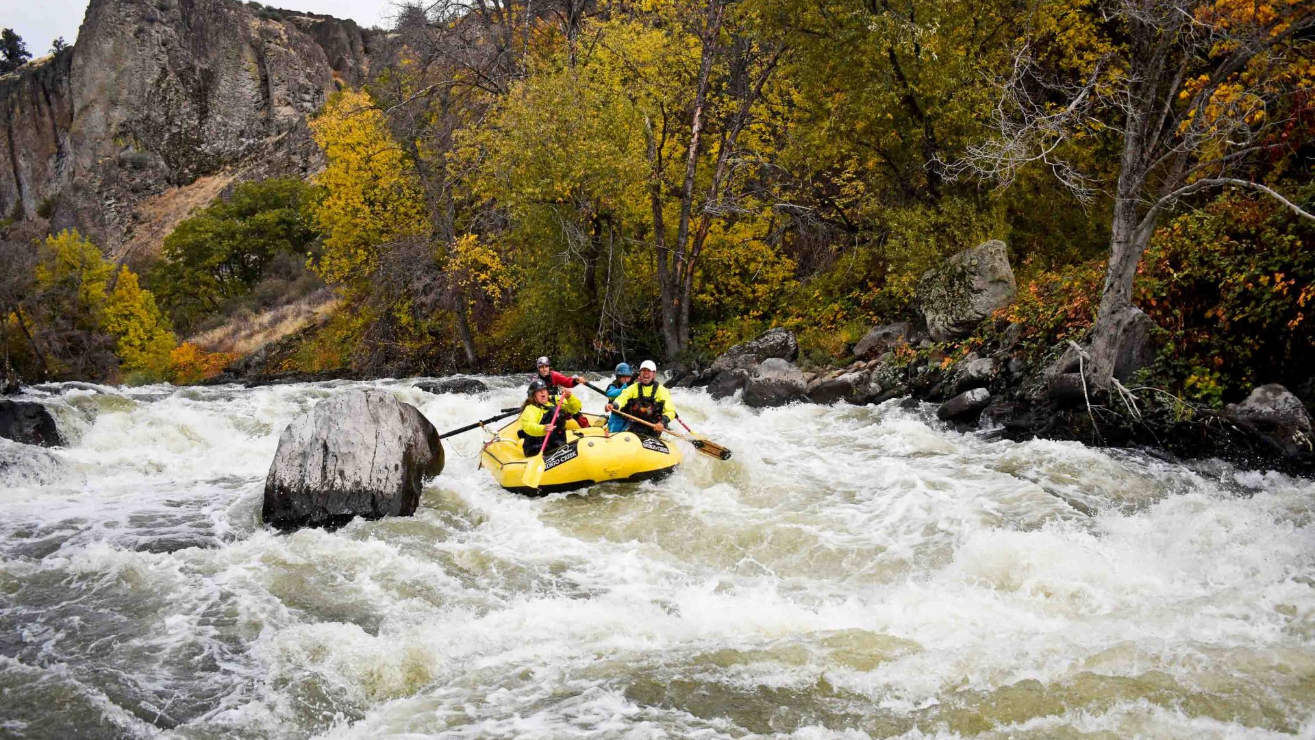 A yellow white water raft goes down a river with tall rocky walls and plants on the edges.