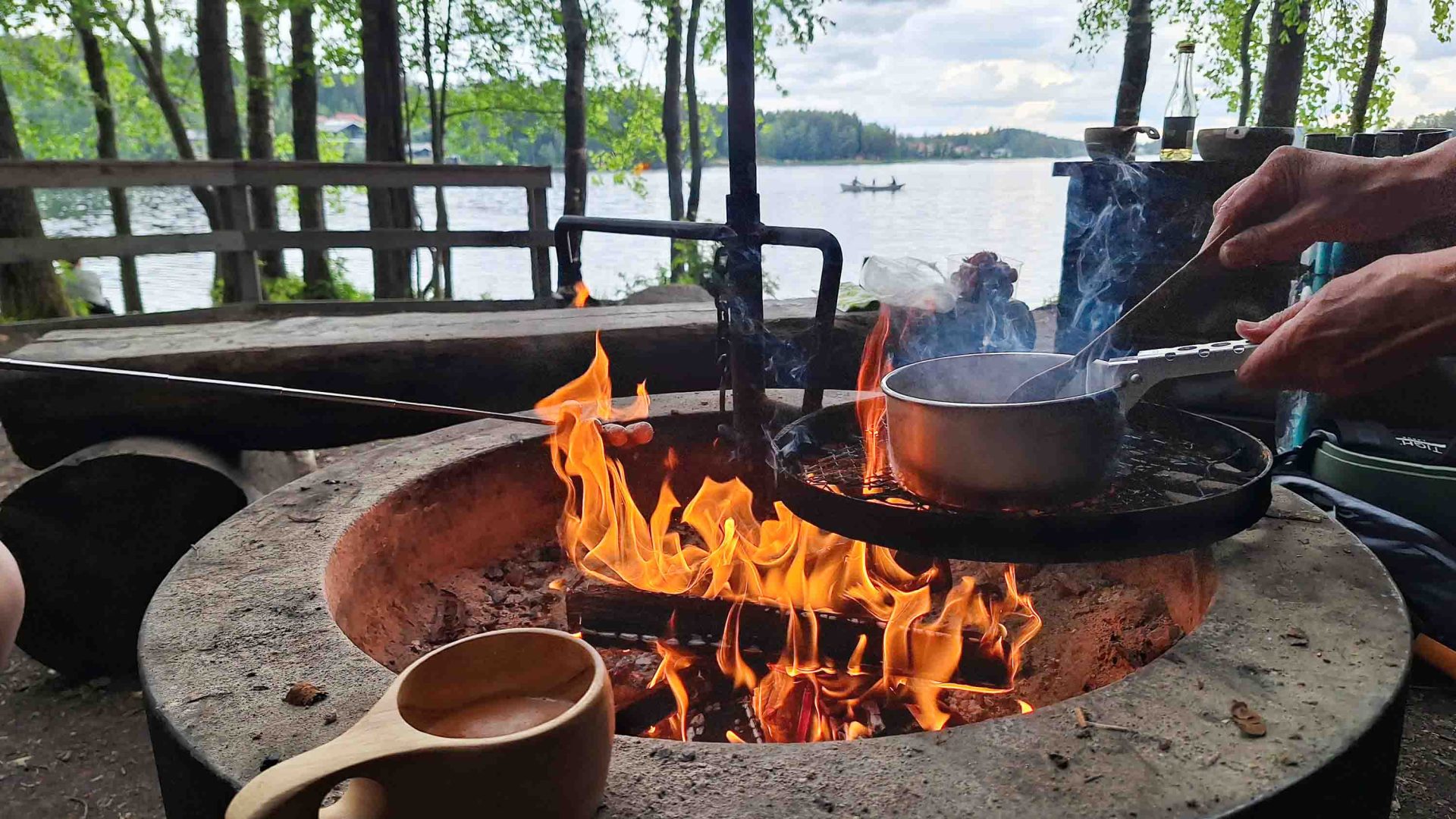 A hand holds a pan over a fire place by a lake.