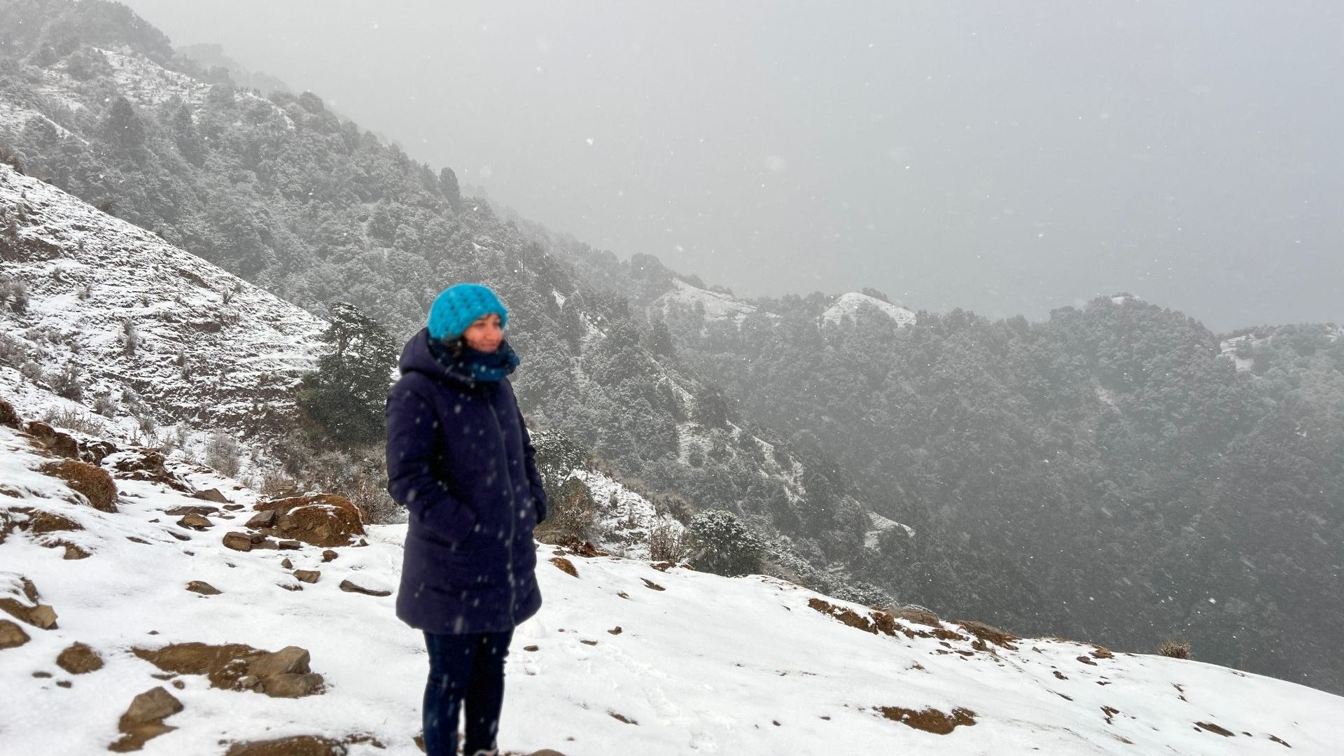 A woman in a blue hat and warm clothes stands against a snowy landscape.