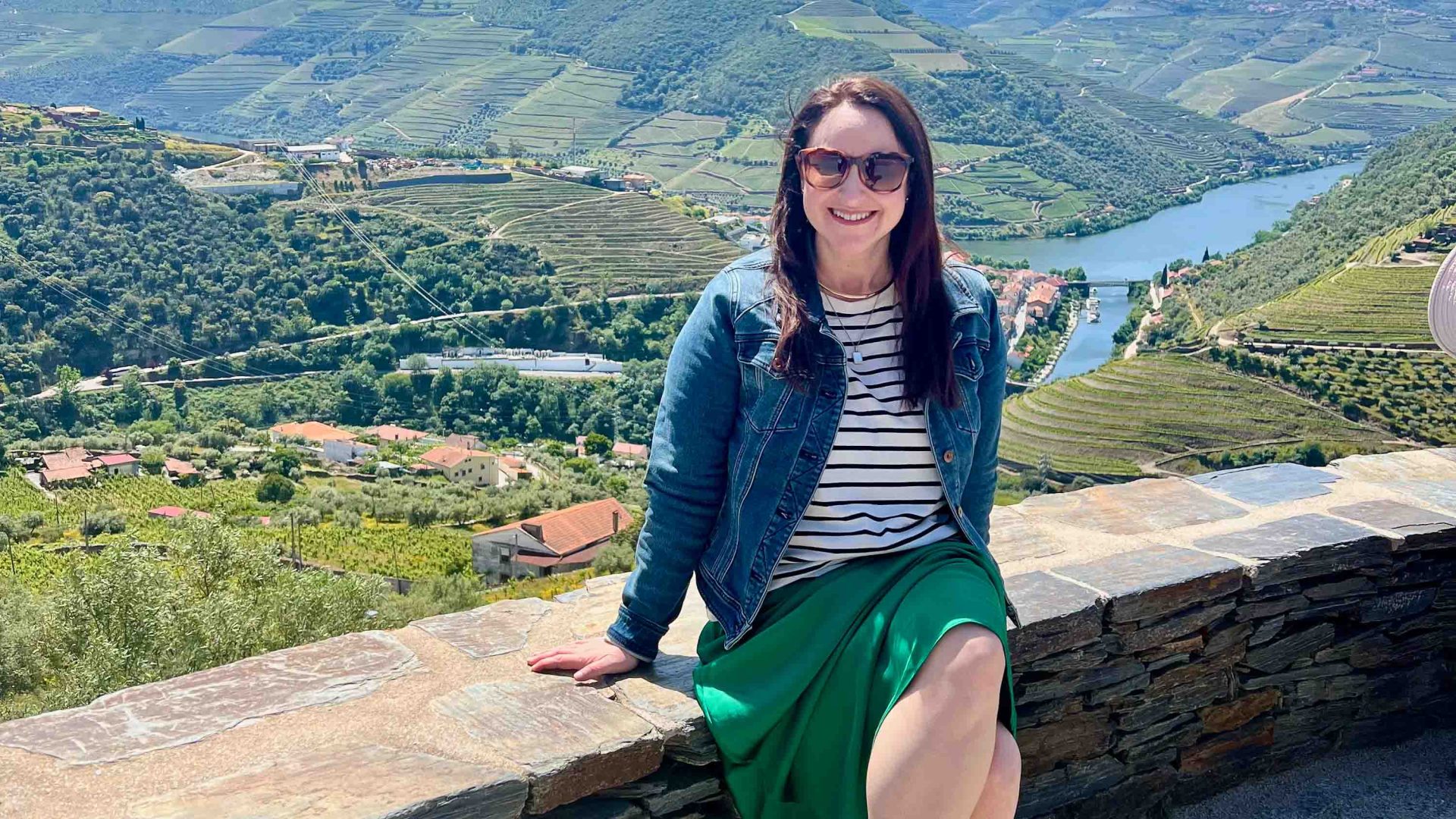 A woman sits on a wall overlooking green fields and a river.