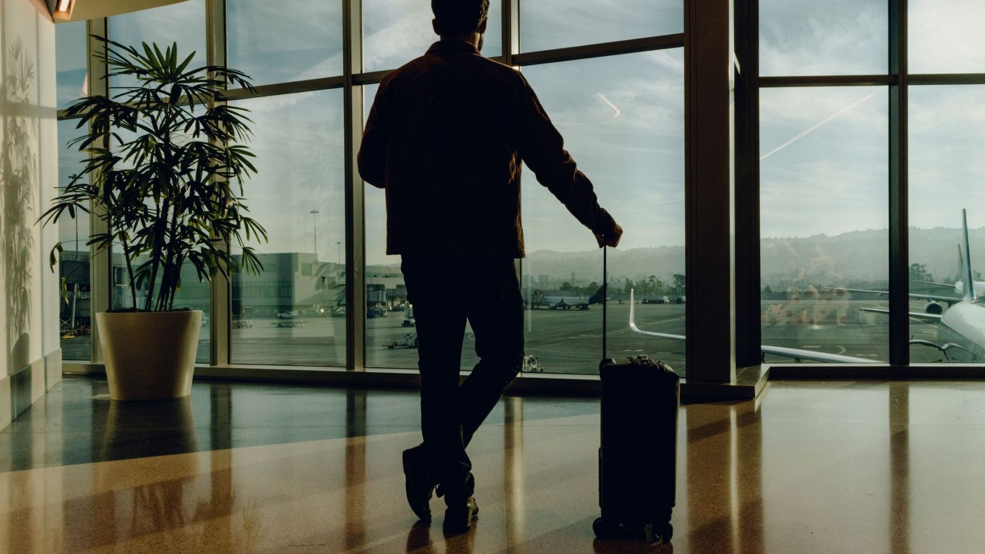 A silhouette of a traveler at an airport, looking away from the camera out a large window. They're holding the handle of a carryon suitcase on wheels.