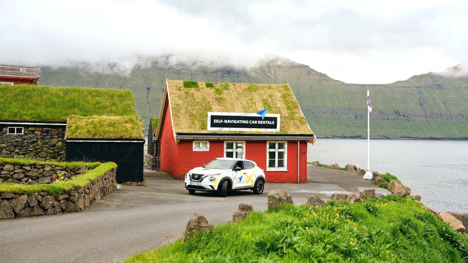 A white rental car in front of a red house with a green turf roof, located at the end of a road overlooking water.